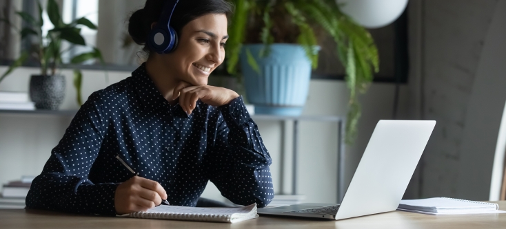 A person sitting and smiling in front of a laptop, wearing headphones and holding a pen to a notepad.